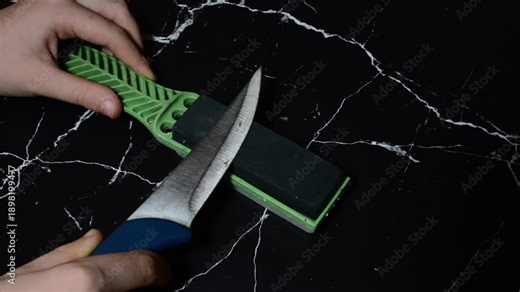 hands sharpening a kitchen knife with a blue handle against a green handheld sharpening stone over a black marble surface.