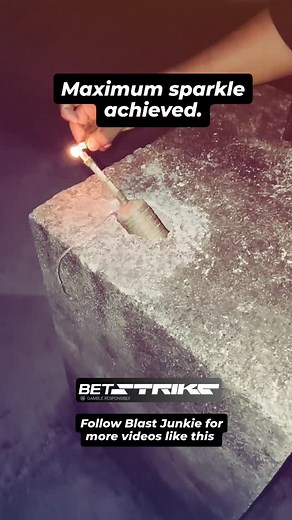 The person is shown carefully igniting the firework's fuse with a handheld sparkler or similar ignition source. After the fuse is lit, the person quickly steps back. ​The firework immediately begins its display, starting with a rapid burst of white and yellow sparks. This quickly intensifies into a powerful, towering fountain of vibrant orange and red glowing embers and sparks. The pyrotechnic blast is quite strong, casting an intense light on the concrete surface before fading out. ​The final s