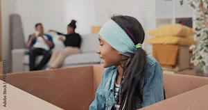 A cute little girl with a hairband sits in a cardboard box used for packing things during a move, sister hides from her brother while they play