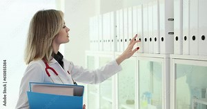 Administrator of medical clinic finds required folder of patient card in rack and checks compliance of data. Medical archive and documentation