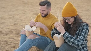 Young Female And Male Friends Eating Take Away Food Sitting On The Beach | Free Stock Video Footage