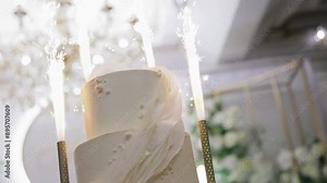 A beautiful birthday cake stands on the table with burning fireworks on the sides. Shooting treats in close-up