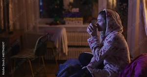 Young woman sitting on bed watching tv and drinking tea