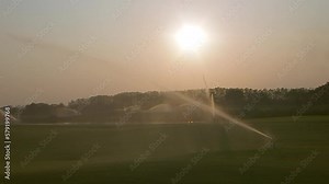 high-pressure sprinkler is spraying water on the golf course grass, spinning in circles to spread water evenly. Sprinklers spraying water against the yellow evening sun at golf course.