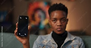 African woman presents cellphone with dark screen in living room, discussing the intuitive design and functionality of mobile interfaces.