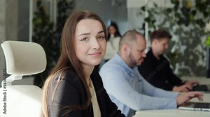 Energetic positive beautiful female programmer works on laptop, writing software for a mobile application. She looks up at the camera and smiles. In the background programmers working alongside her.