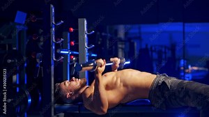 Shirtless young bodybuilder doing bench press for chest. Strong abs. Blue light filter. Video from the side.