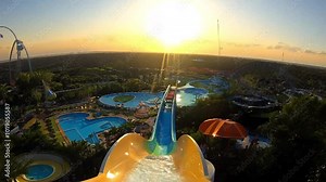 Point-of-view perspective from the top of a water slide at sunset, overlooking a vibrant water park, creating a thrilling and exciting atmosphere
