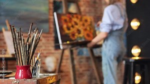 Working process in art studio. Set of painting brushes and palette. Painter creating still life with sunflower on canvas using oil paintings and palette knife. Creative process, relax, leisure, hobby.