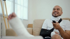 Happy man recovery watching tv using remote from accident fracture broken bone injury with leg splints in cast, neck splints collar, sling support arm. Social security and health insurance concept.