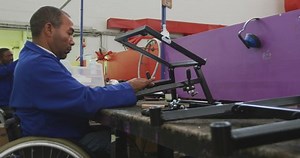 Side view of two disabled African American male workers in a workshop at a factory making wheelchairs, sitting at a workbench assembling parts of a product, both in a wheelchairs, with coworkers