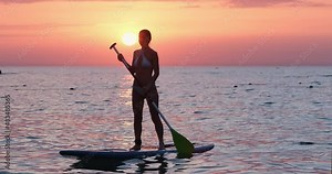 A young girl in a swimsuit floats on a stand up board and holds a balance with an oar, a walk by the sea at sunrise.