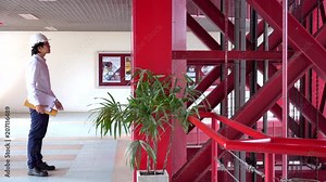Young Asian engineer man or architect with helmet holding architectural drawing and clipboard waiting for lift (elevator) in the red building. Male technician worker working at construction site.