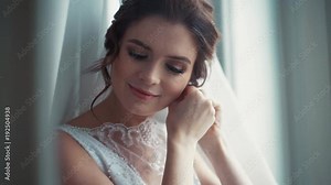 Elegant blue-eyed bride in a beautiful white wedding dress and bridal veil puts on earrings. Wedding preparation, happiness, positive mood