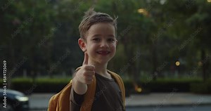 Portrait handsome happy school boy, smiling school boy, little boy with a backpack, standing alone on sunny street outdoors, pretty face primary first grade student. Child shows thumbs up gesture