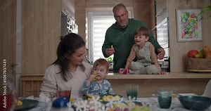 Father and older child interacting joyfully while mother and toddler sit at dining table, warm and playful family moment in a cozy home setting