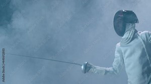 Professional fencer in protective uniform practicing maneuvers with sword alone in dark studio . Fencing demonstration on black smoked background . Shot on Arri Alexa cinema camera in slow motion .
