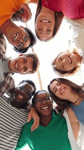 Vertical low angle view of a group of multiracial friends standing on a circle, smiling and embracing together. Young teenagers laughing and looking at camera. Team of people on a coaching meeting
