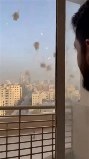 OMG!😲 #staysafe The video opens from a high-rise balcony, overlooking a dense city skyline stretching across the horizon in broad daylight. Apartment buildings fill the foreground and middle distance, while the hazy outline of taller structures rises farther back under a pale sky. A metal balcony railing frames the lower part of the shot, and the atmosphere at first feels tense but still, as if everyone is waiting for something to happen. Almost immediately, the camera captures a dramatic scene