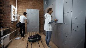 Young women open lockers to put belongings before training. Ladies prepare for sports activity in dressing room with mirrors at fitness studio
