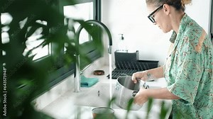 Young adult housewife housekeeper cute woman doing work at hoe washing dishes and pots in white modern kitchen. One female people in indoor house leisure activity. Real lady lifestyle in apartment