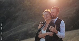 Close-up shot of cute married millennial couple hugging in a deserted spikelet field on warm sunny day. Carring groom wraps his beautiful bride in elegant white wedding dress with jacket. Rest outside