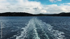 White trail in the Trincomali channel from a ferry sailing on the open sea to vancouver with Salt Spring island on the background on a partly cloudy day. Wide shot