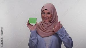 The old lady in a turban, holding a green box in her hand, points to the box.The old lady pointing at the green box draws attention.