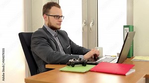 Young, handsome men sitting behind the desk and in using computer, then he is starting play game on pad. He is smiling, positive emotions