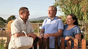 Neighbor conversation. Happy elderly couple chatting friendly with adult man while standing near wooden fence of country house vegetable garden in summer
