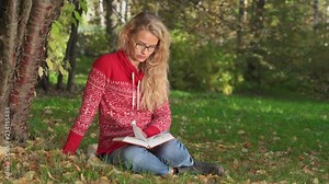 Young woman sitting under a tree in the city park in summer and reading a book. Beautiful girl sitting on green glade under tree branches with book, woman reading novel on nature