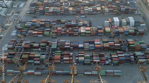 Aerial drone footage showing general container terminal activity at a busy industrial cargo port during evening hours. Stacked shipping containers, cranes and freight logistics infrastructure