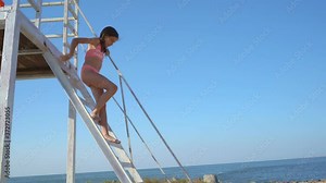 teenage girl goes down stairs lifeguard tower on beach against cloudless sky.