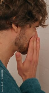 A man in a bathrobe applies lotion to his face while looking in the mirror standing in the bathroom