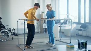 Hospital Physical Therapy Room: Strong Patient with Injury Walking Holding for Parallel Bars, Physiotherapist Encourages Assists, Helps Disabled Person Do Rehabilitative Physiotherapy. Slow Motion