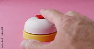 A man's finger is shown pressing down on a small, red button atop a white-and-yellow ring bell on a vibrant pink surface.