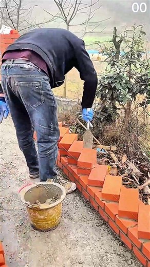 Blue-gloved hands lay red bricks diagonally, mortar secures zigzag wall pattern outdoors