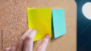 A hand applies a yellow sticky note onto a cork board alongside a blue sticky note, creating a vibrant workspace atmosphere