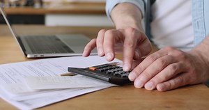 Man use calculator calculates incomes and expenses, pay for loan, entering data results in laptop application, documents, invoices, utility bills lie on table, close up. Manage family budget concept