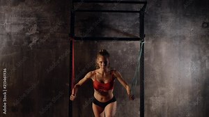 Strong young woman is doing physical exercises for developing muscles of hands, back and abdominal. She is stretching hard rubber ropes, fixed on a rack