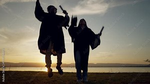 Two graduates are walking on a beach at sunset, holding their caps and gowns. Scene is celebratory and serene, as the graduates are enjoying their graduation day by the water