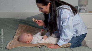 Caring mom playing with her young son holding toy. Baby lies on bed, the woman bent over him. Mother's leisure time with her young child