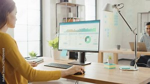 Over the Shoulder: Creative Young Woman Sitting at Her Desk Using Desktop Computer with Screen Showing Project Management, Statistics and Graphs. Office with Diverse Team of Professionals Working