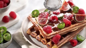 Sifting powdered sugar over Fresh homemade sweet belgian waffles with powdered sugar with fresh raspberries, kiwi berries and figs on white plate