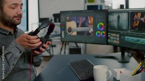 Videographer editing, cuting footage and sound on computer with dual monitors setup putting on headphones working in creative office. Man processing film montage in digital multimedia star up company.