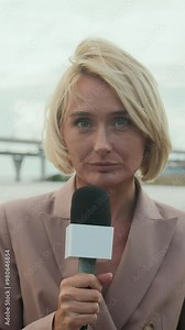 Vertical waist up of Caucasian female reporter standing by waterfront with microphone while delivering news with modern bridge extending over water in background