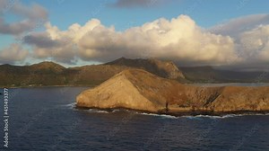 Aerial sunrise view rotating around Rabbit Island, off the coast of Oahu, Hawaii. The island is bathed in golden light with Oahu in background and white puffy clouds in sky.