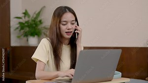 Asian woman working at home, sitting in front computer laptop, hand poked quickly at keyboard, another hand holding mobile phone while talking with friend, smile laughs happily, talked relieve stress.