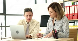Curious colleagues look in laptop and investigation papers comparing data. Excited man and woman sit at table together working on team project