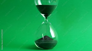 Shot of a sand clock measuring time while the sand is falling down against the green background - old classic timer. Extreme close up of a transparent hourglass with flowing black sand - time concept.